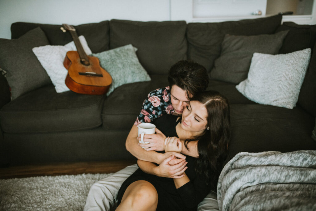 A couple cuddling on a grey sofa, with the man embracing the woman from behind while both hold a mug, a guitar resting in the background, perfect for engagement photo ideas.