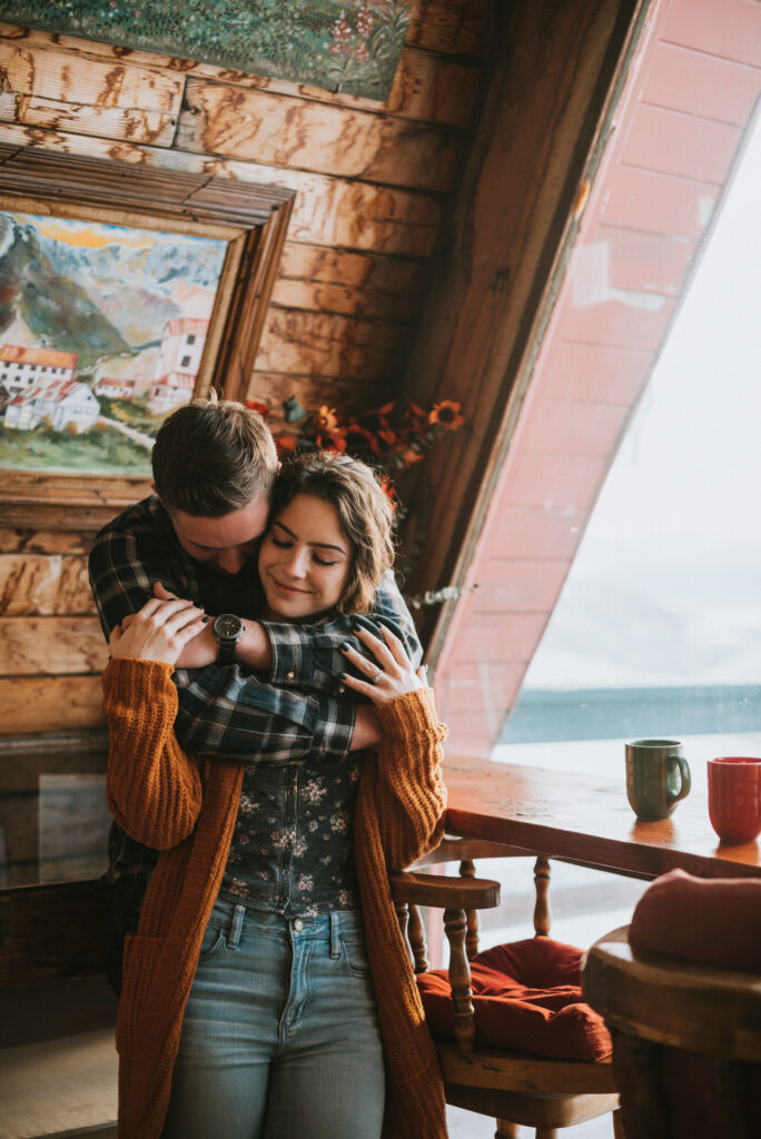 A couple embracing warmly inside a cozy cabin with rustic decor and a scenic window view, perfect for engagement photos.