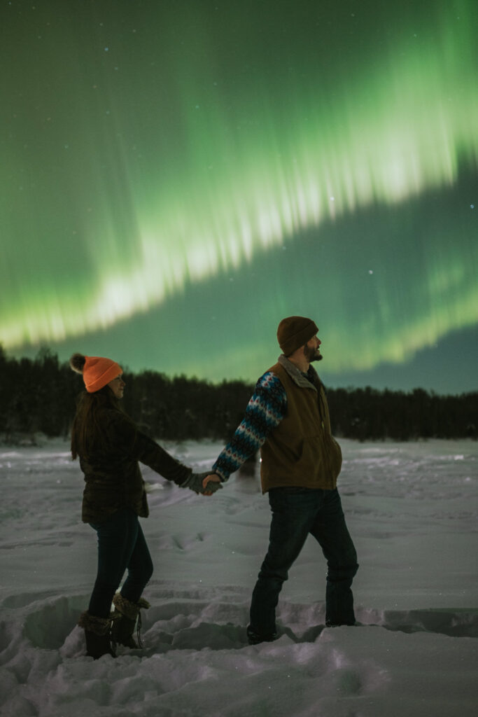 A couple holding hands under the northern lights for their engagement photos on a snow-covered landscape at night.