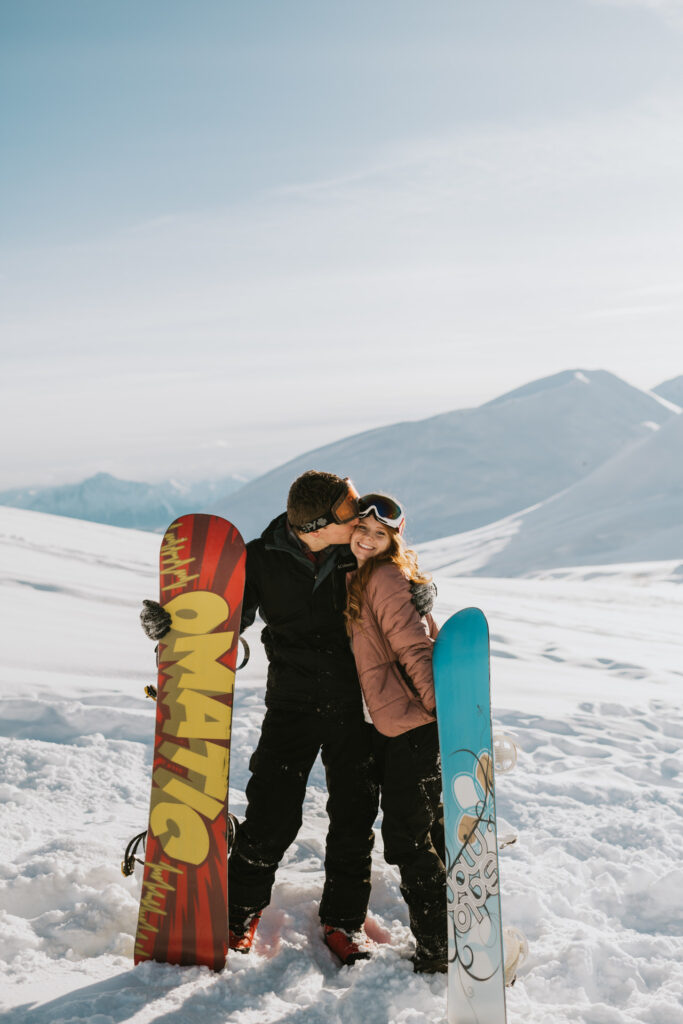 couple standing on a snowy mountainside with their snowboards as he kisses her on the cheek