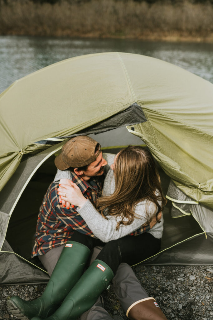 Engaged couple embracing inside a tent beside a tranquil stream during an intimate Alaska engagement session.