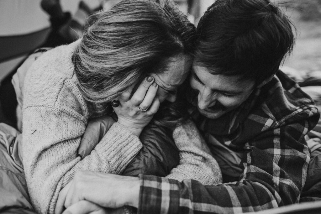 Intimate black and white photo of a couple embracing and laughing together, with one person covering their face shyly, highlighting a genuine, tender moment during a casual outdoor photoshoot in Alaska