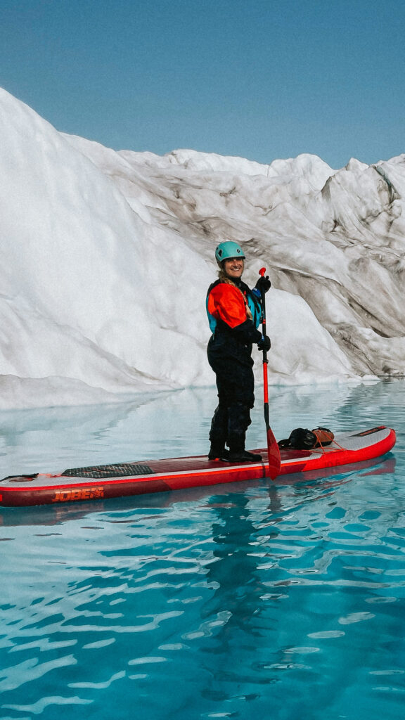 woman in a drysuit standing up on a paddle board while glacier paddling