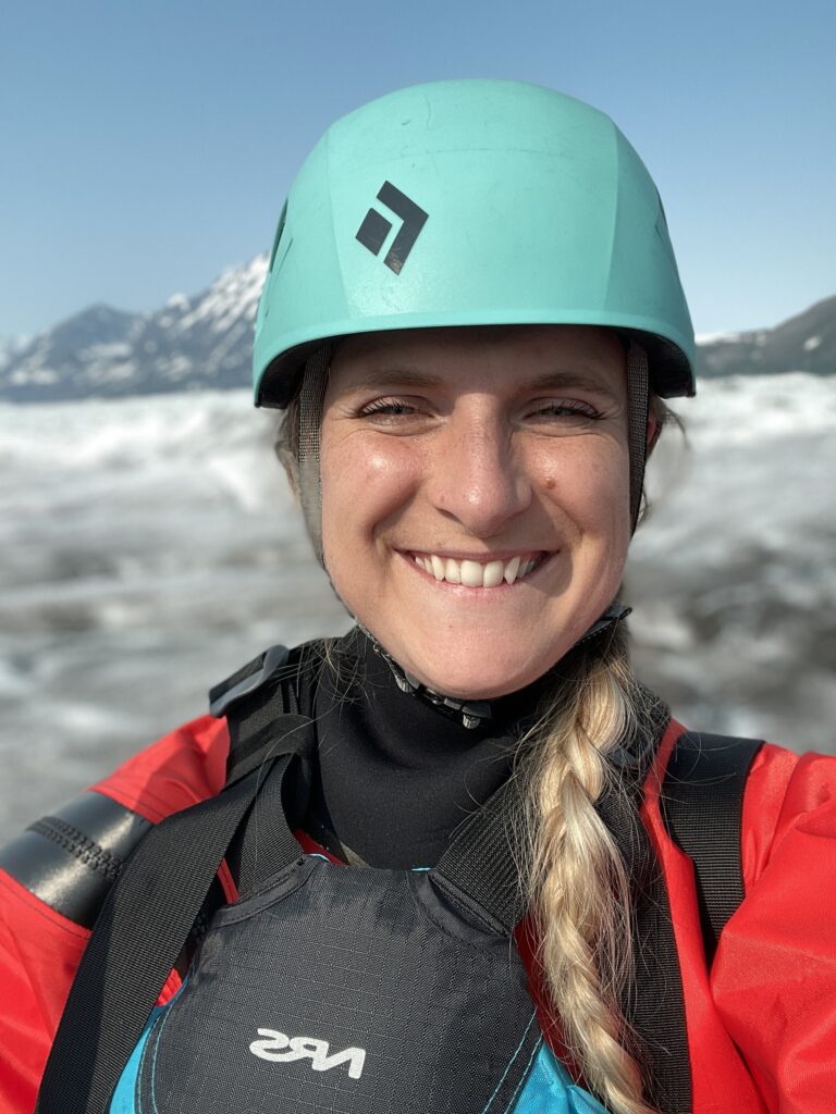 woman smiling at the camera taking a selfie wearing a dry suit and helmet while glacier paddle boarding