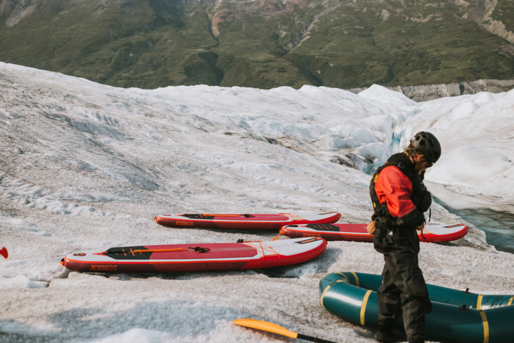 A person in cold weather gear standing near several paddleboards on a glacier, with green mountains in the background, ready for glacier paddle boarding.
