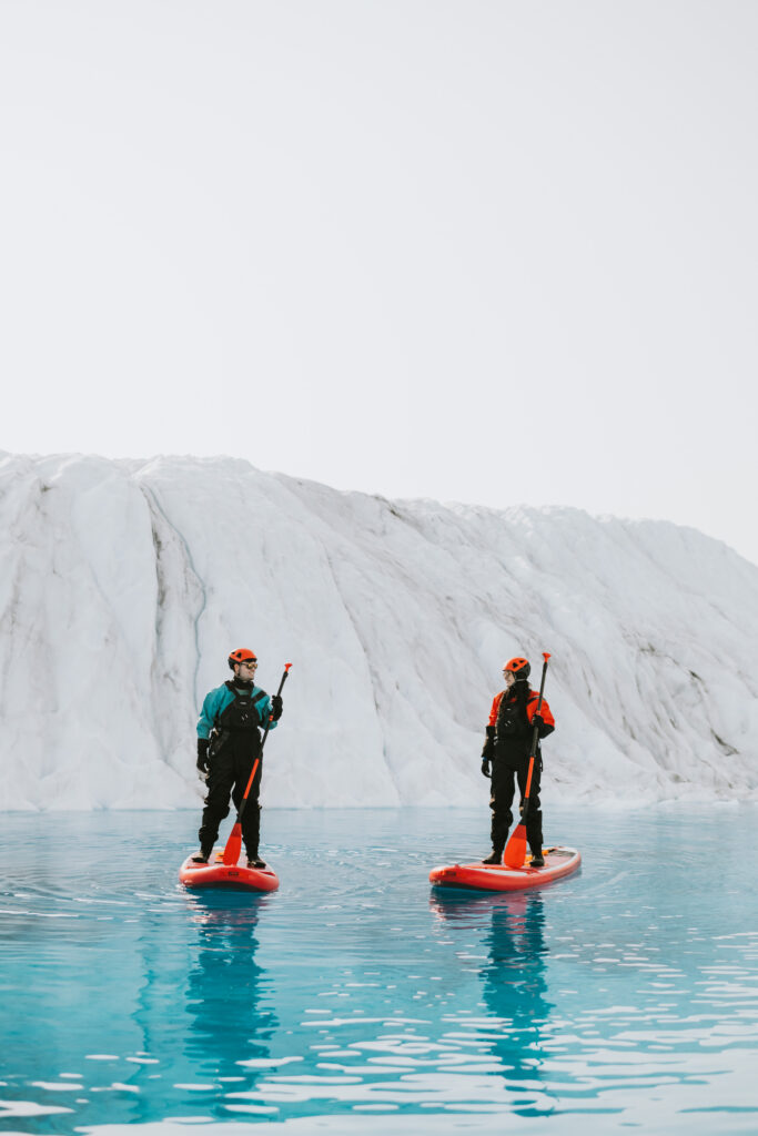 Two people glacier paddleboarding on a large, icy glacier in calm, reflective blue waters.