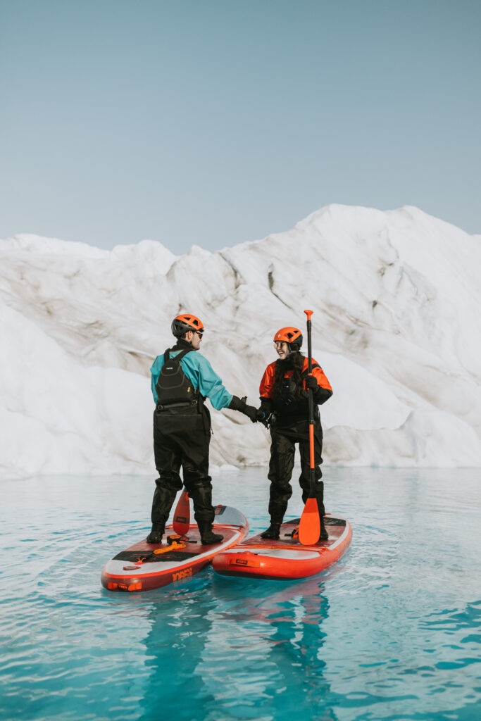 Two people on stand-up paddleboards holding hands on a glacier lake while glacier paddle boarding, surrounded by icy cliffs under a clear blue sky.