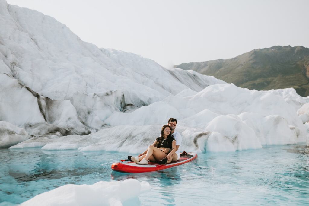 A couple sits smiling on a paddle board surrounded by icy glacier formations in clear blue waters under a bright sky.