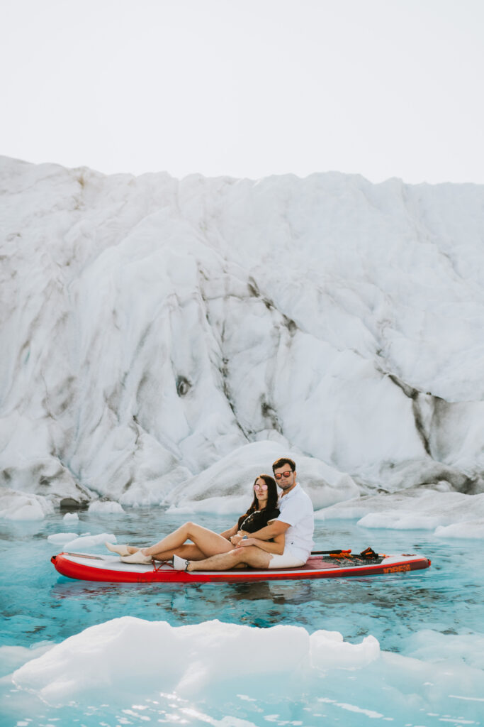A couple sits on a red paddleboard on a glacial lake, surrounded by ice and a stunning glacier backdrop, enjoying glacier paddle boarding.