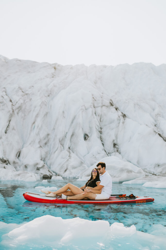 A couple sits closely on a red kayak surrounded by icy waters against a backdrop of a white glacier