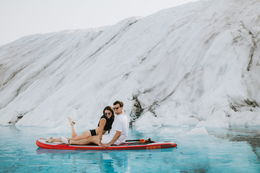 A couple sits on a red kayak in front of an ice wall, surrounded by icy waters as they prepare paddle board.