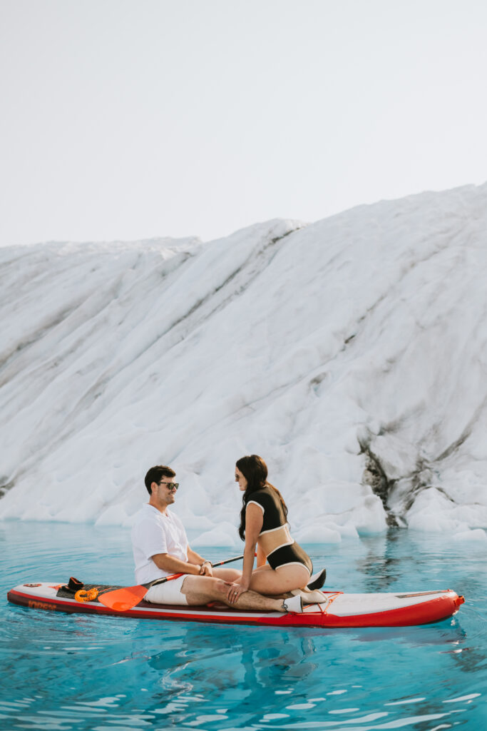 A man and a woman paddle boarding in clear blue waters in front of a white ice wall on a glacier.