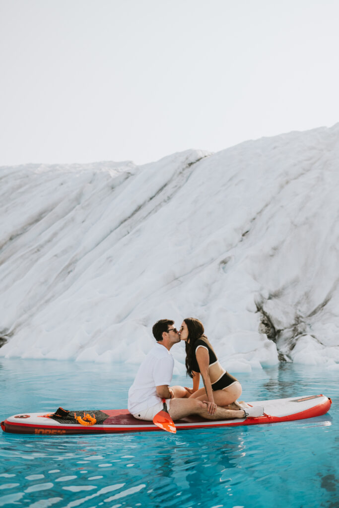 A couple kissing on a red paddleboard in front of a glacier, surrounded by icy blue waters while enjoying glacier paddle boarding.