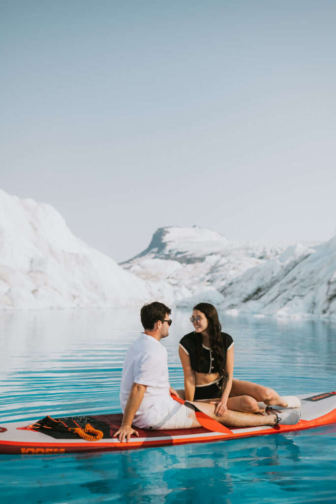 A couple sitting on a glacier paddle board, engaging in conversation on a clear blue glacial lake, with ice formations and mountains in the background.