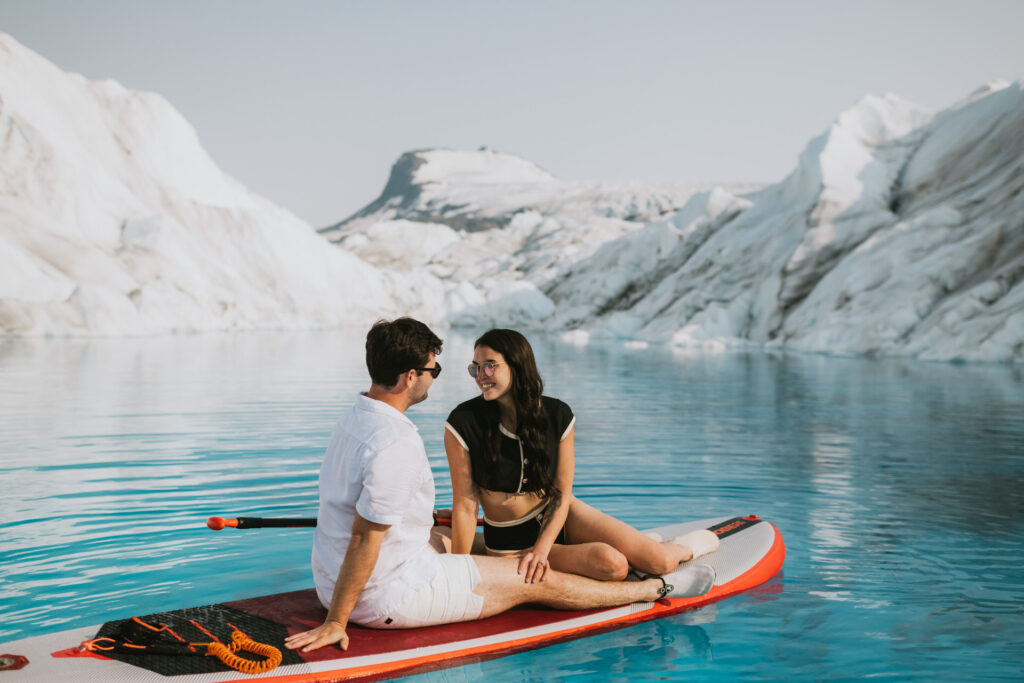 A couple enjoys glacier paddle boarding in calm, blue waters, surrounded by ice formations and engaging in conversation.