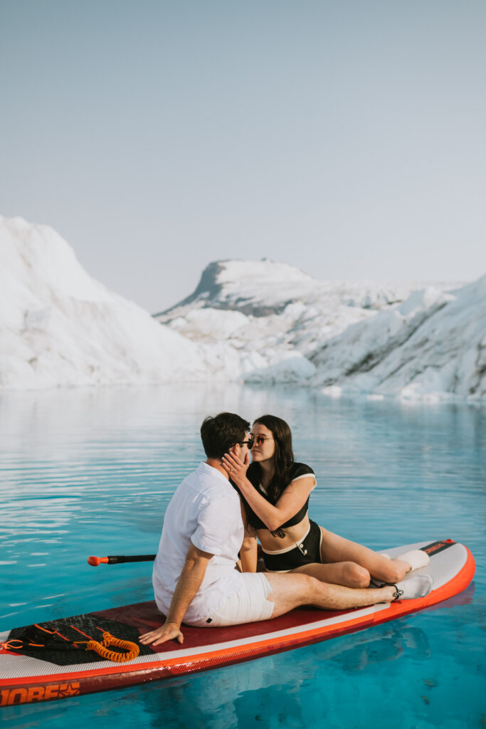 A couple kisses while glacier paddle boarding in clear blue waters against a backdrop of glossy white glaciers.