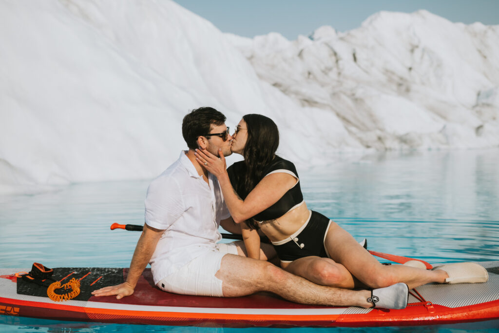 A couple kissing while glacier paddle boarding in a serene, icy water setting with prominent white glaciers in the background.