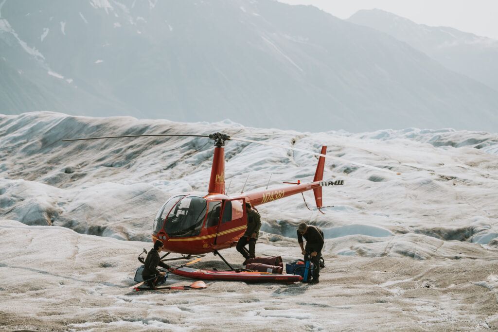 A red helicopter landed on a rugged glacier with two people preparing glacier paddle boarding gear surrounded by mountains.