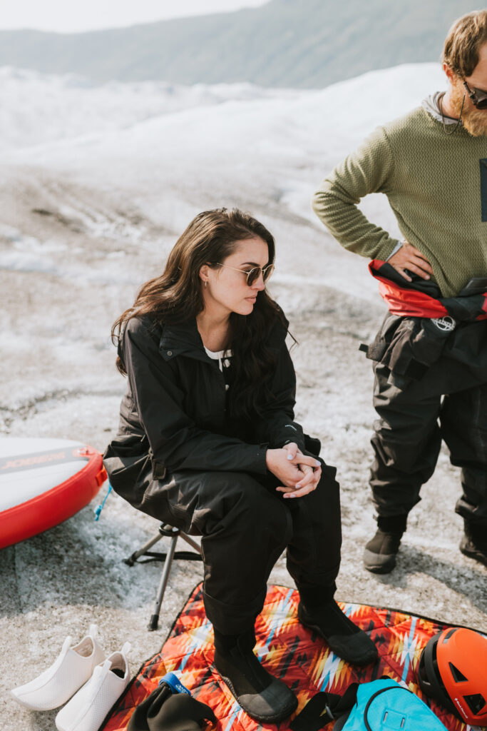 A woman in sunglasses sits on a folding chair in a dry suit by a paddle board on a glacier with a man standing beside her.