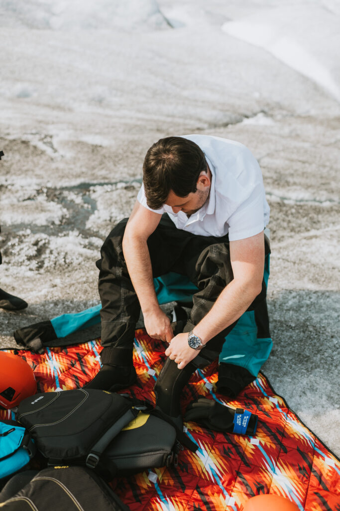 Man in a white shirt putting on black water boots while sitting on a colorful mat with glacier paddle boarding gear around him.