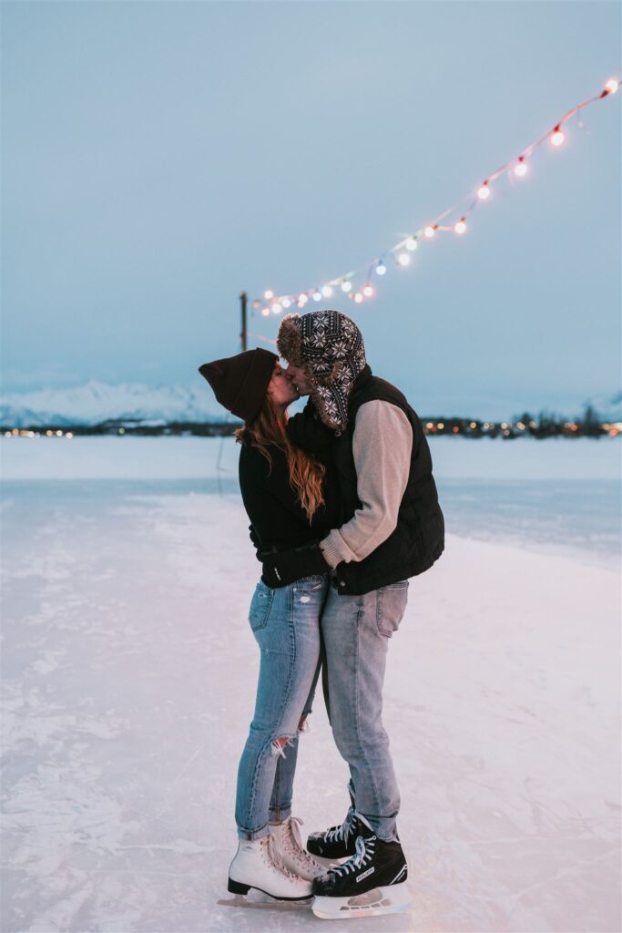 Couple sharing a kiss on an ice skating rink under twinkling string lights, with a serene, snow-covered Alaskan landscape in the background, capturing a romantic winter engagement session.