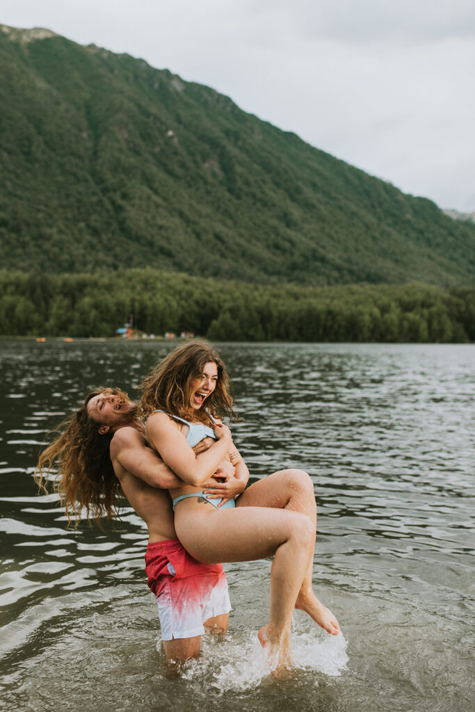 Happy couple laughing while playfully embracing in a picturesque Alaskan lake, with lush mountains in the background, capturing a spontaneous moment during their engagement session.