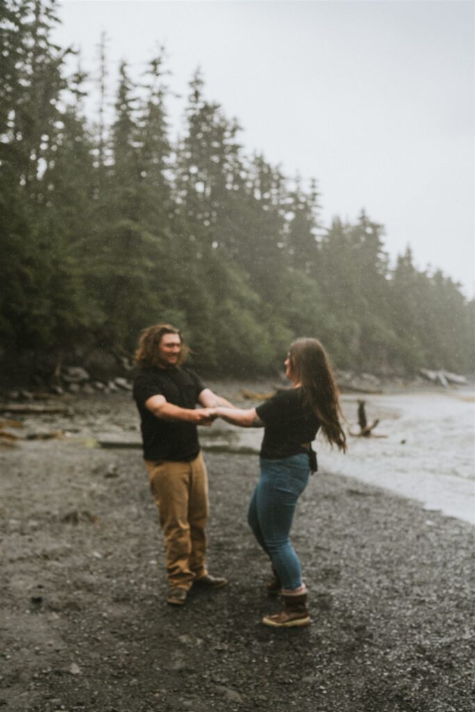 A couple holds hands and smiles on a misty, forested beach. They are casually dressed, and the shoreline is visible in the background.