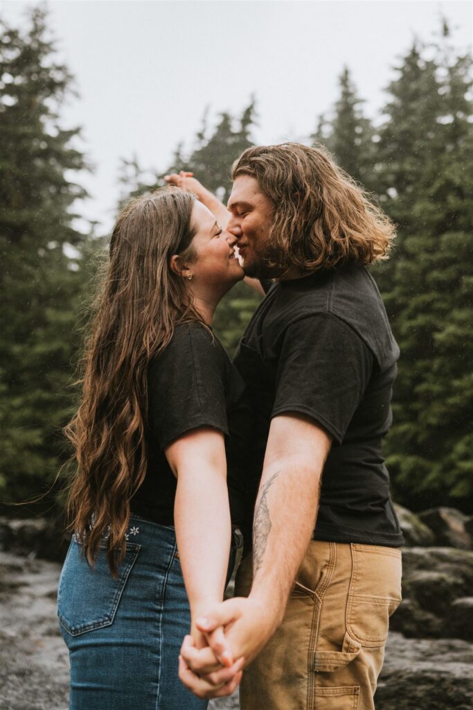 A couple stands close together outdoors, holding hands and leaning in for a kiss, with a backdrop of trees.