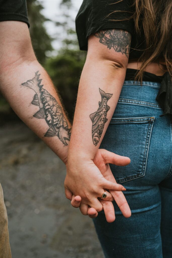 Two people holding hands, each with fish tattoos on their arms. The background shows a blurred natural setting.