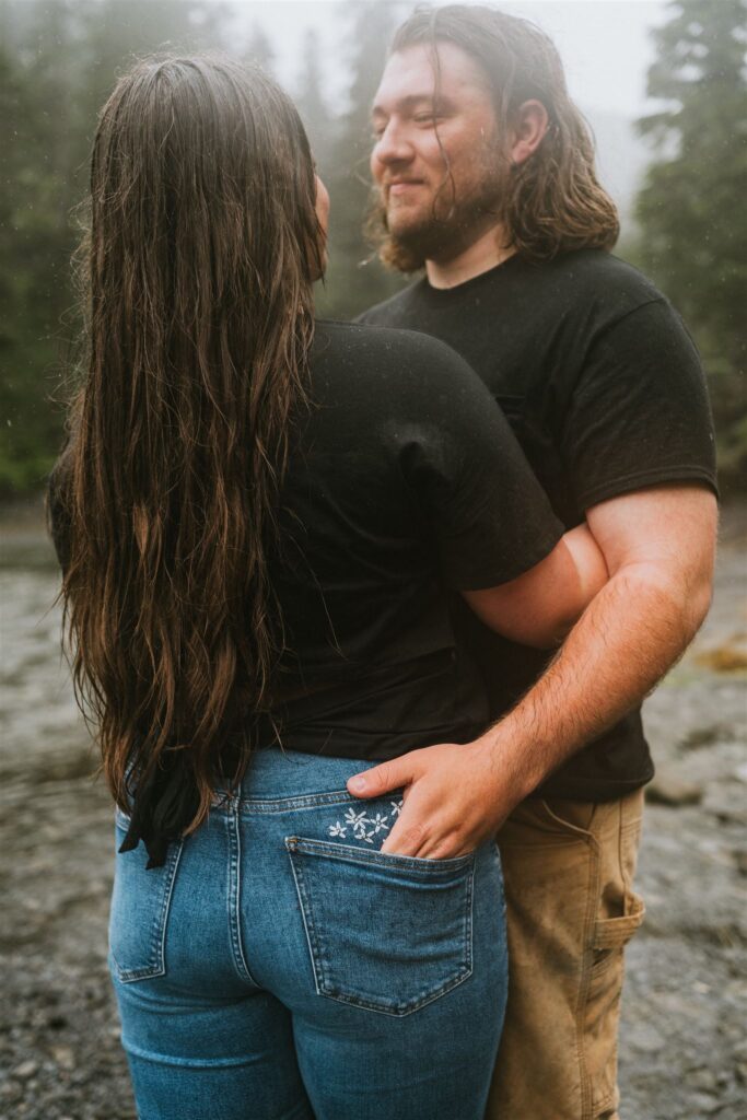 A couple stands together outdoors, with the man's hand resting on the woman's back pocket. They are both wearing casual clothes.