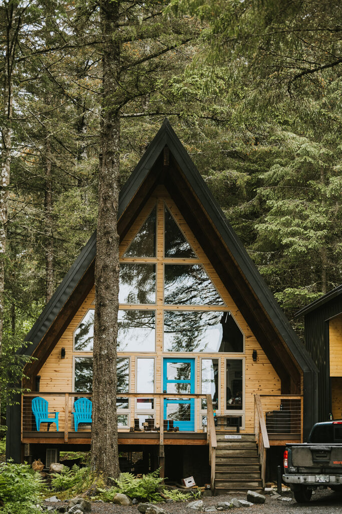 A-frame cabin surrounded by trees, with large glass windows and a blue front door. Two blue chairs sit on the porch. A black pickup truck is parked in front.