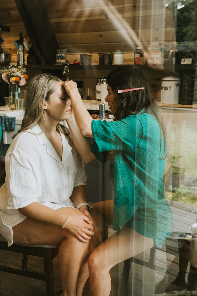 A woman in a green shirt applies makeup to another woman seated in front of her, as seen through a window, with a cozy room in the background.