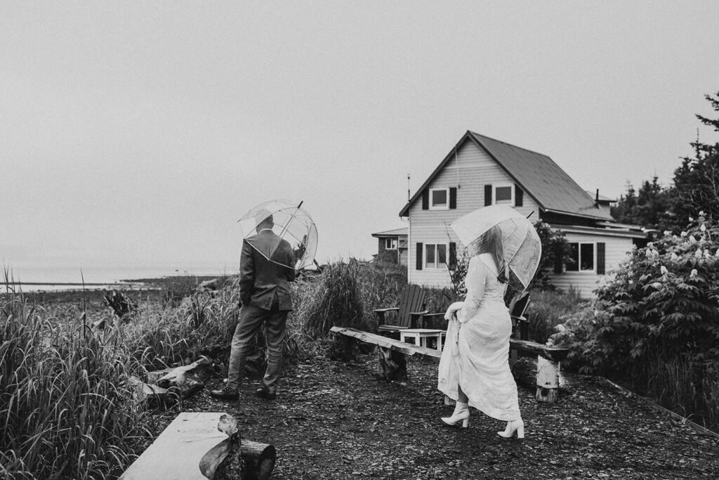 A couple with clear umbrellas walk on a path near a coastal house on a cloudy day.