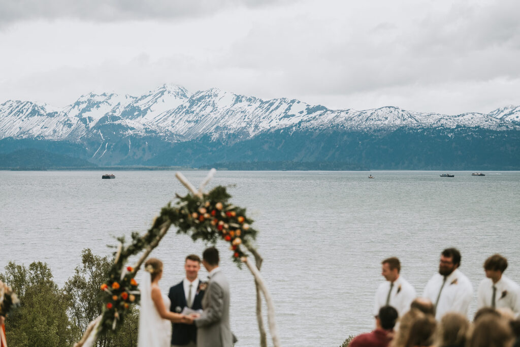 A wedding ceremony by a lake with snowy mountains in the background. An arch decorated with flowers frames the couple, and several people stand nearby.