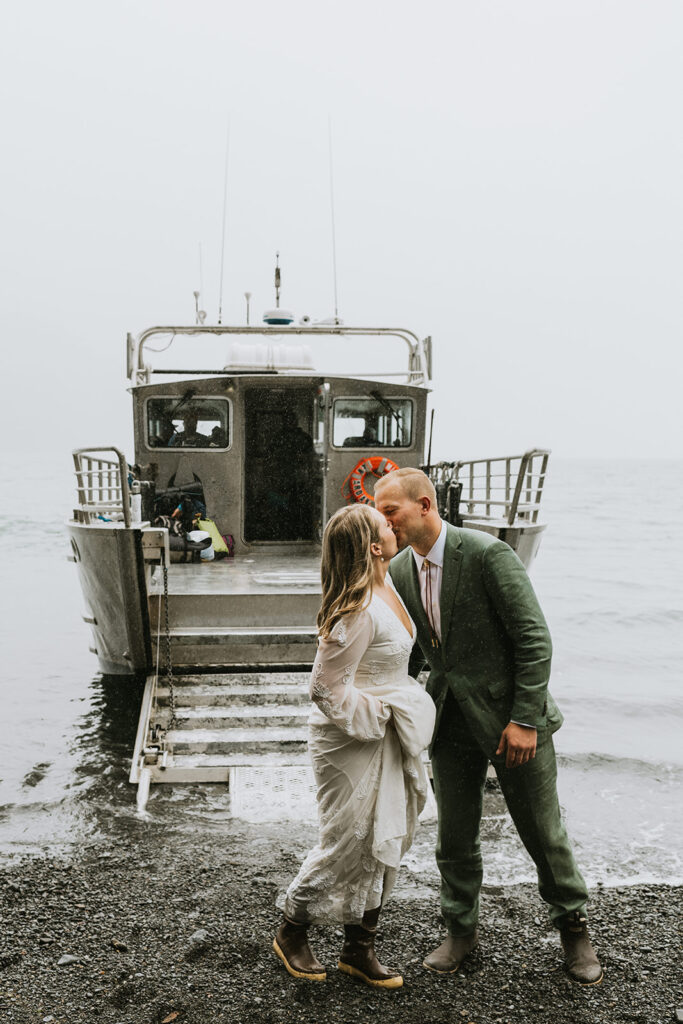 A couple in formal attire kisses on the shore in front of a docked boat on a foggy day.