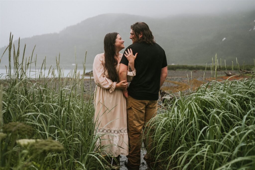 A couple stands close together, surrounded by tall grass, with misty mountains in the background.