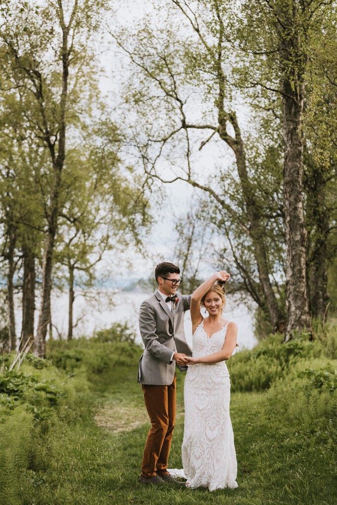 A couple dances outdoors on a grassy path surrounded by trees, with a lake in the background. The woman wears a white dress, and the man wears a suit with a bow tie.