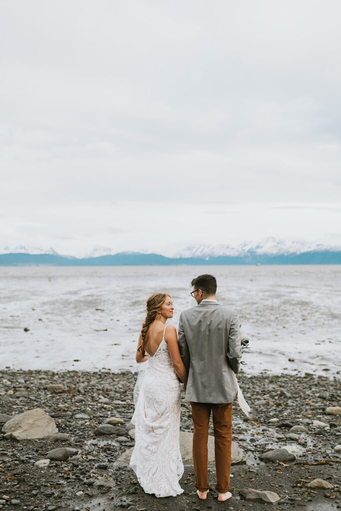 A couple in wedding attire stands on a rocky beach, facing the ocean with snowy mountains in the background.