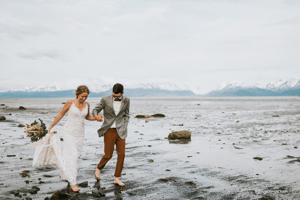 A couple, dressed in wedding attire, walk barefoot along a rocky beach with mountains in the background.