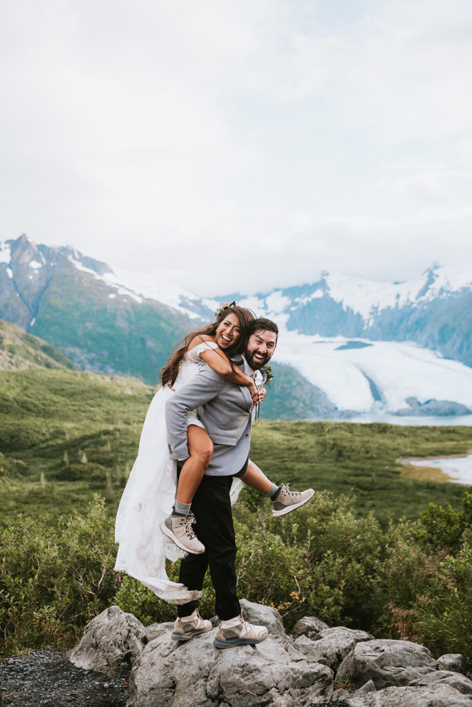 A woman piggybacks on a smiling man, both dressed casually. They stand on rocks with a scenic backdrop of mountains and a glacier.