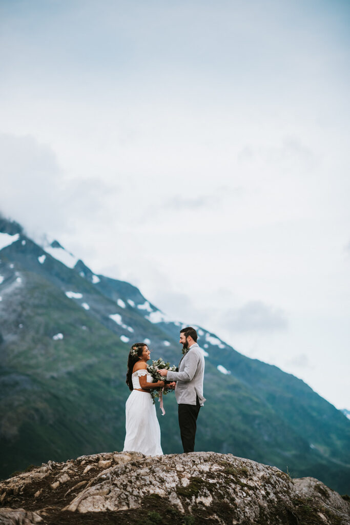 A couple stands on a mountain peak, the bride holding a bouquet, with snow-capped mountains in the background under a cloudy sky.