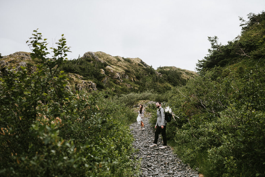 A couple walks along a rocky path through lush greenery and hills, with overcast skies above.