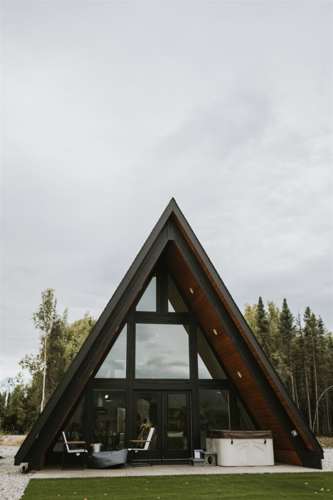 A-frame cabin with large glass windows, nestled among Alaska's stunning trees. Under the overcast sky, relax on the patio in cozy chairs or soak in the hot tub after a day's adventure. Perfect for those who travel to Alaska seeking tranquility and breathtaking landscapes.