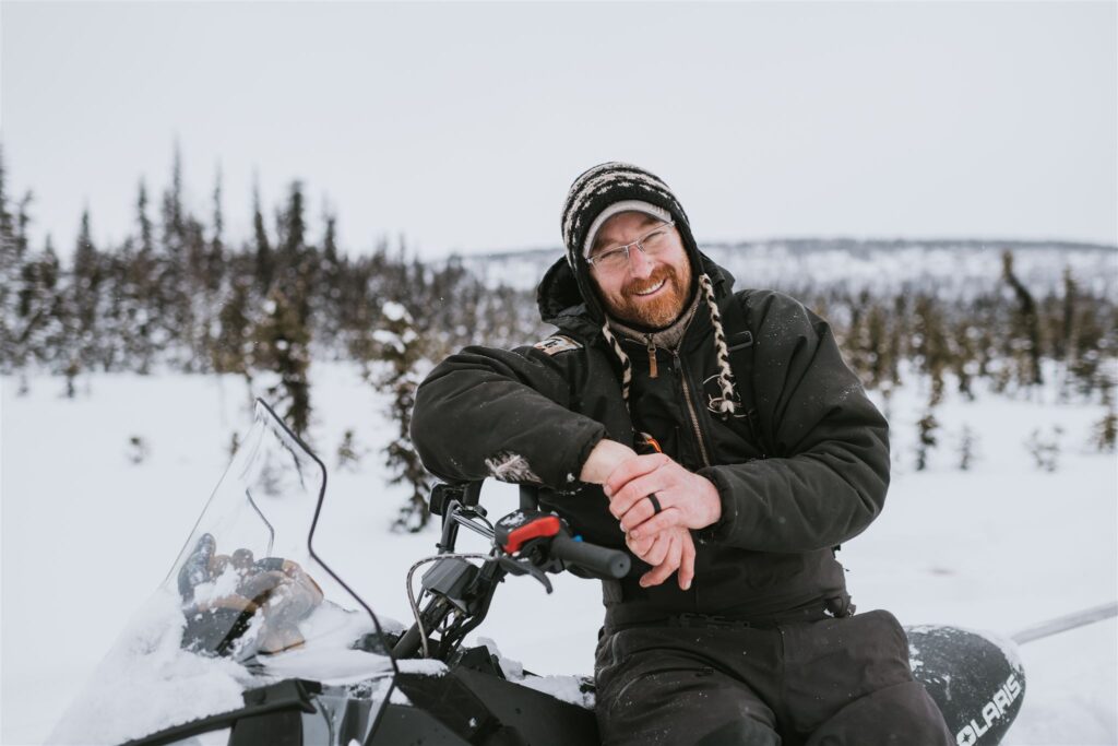 Bundled in winter clothing, a person embarks on an adventurous journey by snowmobile through Alaska's snowy landscape, with a backdrop of majestic trees.