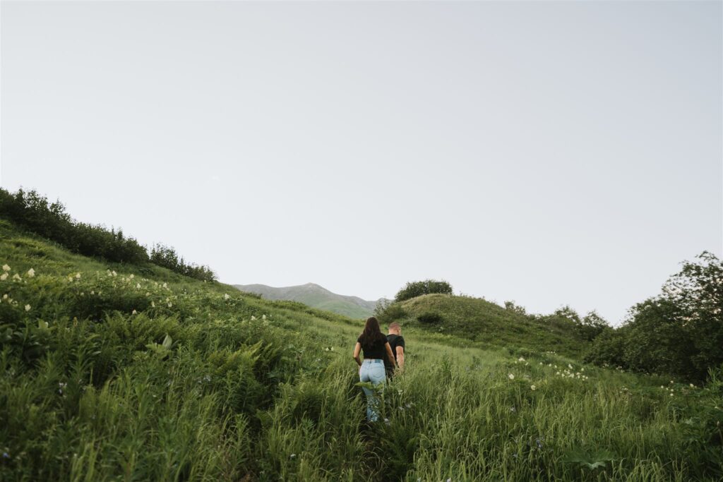 Two people walking up a grassy hill surrounded by lush green foliage under a clear sky, reminiscent of a serene journey you might experience if you travel to Alaska.