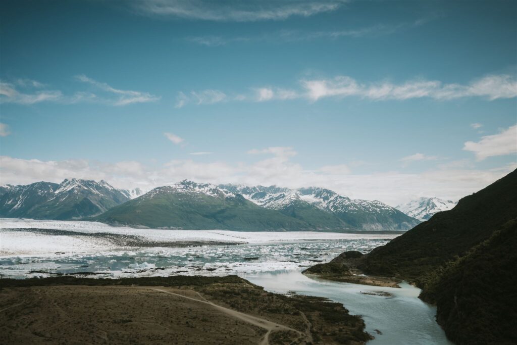 Scenic view of a river flowing through a valley with snow-capped mountains in the background under a clear blue sky, inviting you to travel to Alaska and experience nature's untouched beauty.