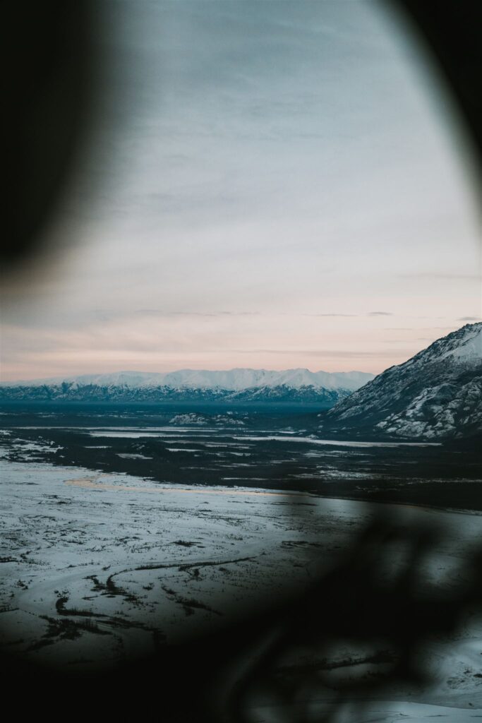 Through the airplane window, the majestic view unfolds—snow-covered mountains and plains stretch under a cloudy sky, inviting thoughts of travel to Alaska.