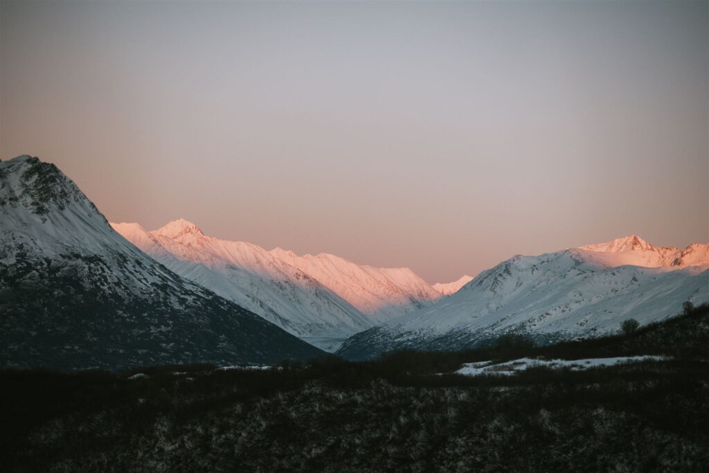 Snow-covered mountain range with a pinkish sunset glow on the peaks under a clear sky, inviting you to travel to Alaska and experience nature's breathtaking beauty firsthand.