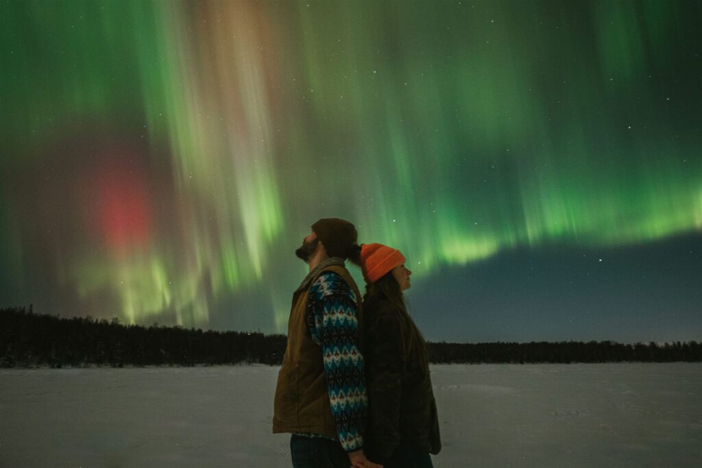 Bundled in cozy winter clothing, two people stand on a snowy Alaskan landscape, captivated by the vivid display of the northern lights dancing across the night sky—a travel to Alaska that promises an enchanting celestial experience.