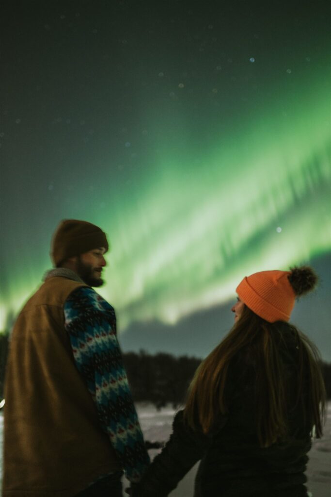 Bundled in winter clothing, a couple stands beneath the shimmering green Northern Lights, dreaming of their next adventure. Inspired by this breathtaking spectacle, they consider a journey to travel to Alaska and experience its untouched wilderness firsthand.